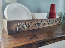 Load image into Gallery viewer, A rustic dark wood sign or tray labeled 'plates napkins elbow cups' in raised silver script, styled on a wooden table with white paper plates, folded napkins, and red cups in a home setting.