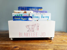 Load image into Gallery viewer, A white wooden children's book box with small caster wheels, decorated with a pink elephant illustration and the personalized name 'BLAKELEE JO' in pink lettering. The box is filled with children's books and sits on a wooden table against a gray wall.