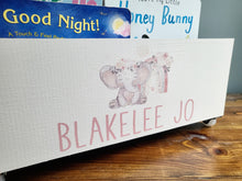 Load image into Gallery viewer, A white wooden bookshelf or bookcase with a painted pink elephant illustration and the personalized name 'BLAKELEE JO' in pink lettering, shown on a wooden tabletop with children's books visible behind it.