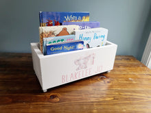 Load image into Gallery viewer, A white wooden children's book box with pink personalized text reading 'BLAKELEE JO' and a pink elephant illustration, filled with colorful children's books, placed on a wooden table against a blue-gray wall.