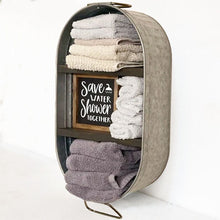 Load image into Gallery viewer, A wall-mounted oval metal storage organizer filled with folded towels and a small framed sign that reads 'Save Water Shower Together.'.