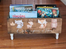 Load image into Gallery viewer, A rustic wooden children's book bin with white painted legs, decorated with cute woodland animal illustrations (raccoon, deer, fox, bear) and the phrase 'Once upon a time.' It is filled with children's books and sits on a wooden floor.
