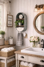 Load image into Gallery viewer, A rustic farmhouse-style bathroom scene featuring a gray metal wall-mounted shelf with a clock, small plant, folded towels, and a hanging gray towel. A white sink with a wood countertop, wicker baskets of rolled towels, a round wood-framed mirror, and a window with sheer curtains are also visible.