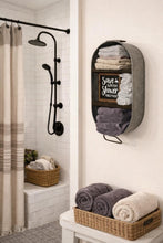 Load image into Gallery viewer, A styled bathroom scene showing a gray fabric wall-mounted towel organizer holding folded towels and a small chalkboard sign, with additional towels in woven baskets on a bench and near a shower area.