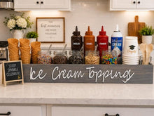 Load image into Gallery viewer, A rustic kitchen-themed ice cream topping bar setup with a large gray wooden sign reading 'Ice Cream Toppings' on a white countertop. Visible ingredients include chocolate, caramel, strawberry, and whipped cream bottles, jars of sprinkles and nuts, waffle cones, cups, and wooden serving utensils.