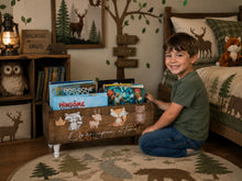 Load image into Gallery viewer, A smiling young boy kneels on a round forest-themed rug next to a rustic wooden storage bin filled with children's books in a cozy, decorated bedroom.