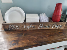 Load image into Gallery viewer, A rustic dark wood sign or organizer tray labeled 'plates napkins silverware cups' holding white paper plates, white napkins, white plastic forks, and a stack of red plastic cups on a wooden table.
