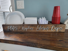 Load image into Gallery viewer, A rustic wooden sign with cursive text reading 'plates napkins silverware cups' placed on a dark wood counter, holding white paper plates, white napkins, white plastic forks, and a stack of red plastic cups.
