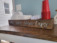 Load image into Gallery viewer, A rustic dark wood sign engraved with the words 'plates napkins silverware cups' in white script, styled on a wooden counter holding red plastic cups, white paper plates, and white napkins.
