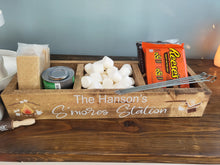 Load image into Gallery viewer, A rustic wooden s'mores-themed tray labeled 'The Hanson's S'mores Station' filled with marshmallows, a can of coffee, a bag of Reese's, graham crackers, and skewers on a wooden table.
