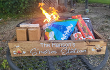 Load image into Gallery viewer, A rustic wooden crate labeled 'The Hanson's S'mores Station' sits outdoors on a patio table, with fire burning behind it and assorted snack packages and wrapped treats arranged inside. Visible treats include a blue bag of Lay's Puffed Rice and candy bars such as Reese's and Kit Kat.
