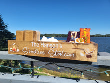 Load image into Gallery viewer, A rustic wooden gift basket tray labeled 'The Hanson's S'mores Station' filled with graham crackers, marshmallows, and snack packages, set outdoors by a lake with trees and blue sky in the background.
