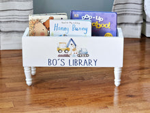 Load image into Gallery viewer, A white wooden children's book bin with turned legs, filled with books, on a hardwood floor. The front of the bin is painted white and features the text 'BO'S LIBRARY' with a small illustration of construction vehicles.
