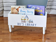 Load image into Gallery viewer, A white wooden children's book box with legs, labeled 'BO'S LIBRARY' and decorated with construction vehicle illustrations, filled with children's board books on a hardwood floor.
