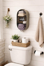 Load image into Gallery viewer, A wall-mounted fabric towel and soap dispenser organizer in a rustic gray-brown color, holding folded towels and a small chalkboard sign that reads 'wash'. The organizer is mounted on a white shiplap bathroom wall above a toilet with a wicker basket and plants on top, and a beige towel hangs from a black hook on the right.

