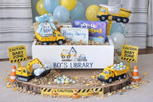 Load image into Gallery viewer, A baby shower themed construction boy gift box display on a wood slice base, decorated with toy construction vehicles, caution signs, balloons, and a 'BO'S LIBRARY' book box.
