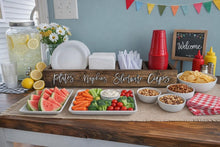 Load image into Gallery viewer, A styled party setup on a wooden table with a large glass jar of lemonade, paper plates, napkins, elbowware cups, bowls of fruit, vegetables, chips, and snacks, plus a small welcome sign and bunting against a blue wall.
