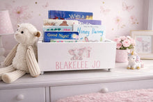 Load image into Gallery viewer, A white wooden children's book box with pink personalized text reading 'BLAKELEE JO' and pink elephant illustrations, styled in a nursery setting on a dresser next to a plush teddy bear and a small unicorn figurine.
