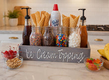 Load image into Gallery viewer, A wooden 'Ice Cream Toppings' serving tray filled with glass bottles of syrups, jars of sprinkles, chocolate chips, candy, marshmallows, and waffle cones, styled on a kitchen counter.
