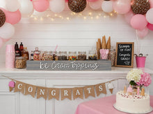 Load image into Gallery viewer, A decorated ice cream topping bar setup with pink and white balloons, string lights, jars of toppings, pink cups and napkins, a chalkboard sign reading 'Scoops of FUN! Congrats! Grad!', a burlap 'CONGRATS' banner, and a white cake on a pink tablecloth.
